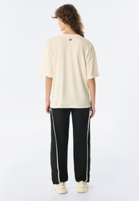 Crème jersey T-shirt met een losse pasvorm, korte mouwen, en logo op de rug; gecombineerd met zwarte broek met witte zijstrepen en sneakers.