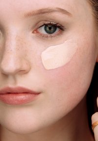 Lys beige foundation på en lys hudtone, som viser en glat tekstur og synlige fregner på huden. Subtil leppestift.
