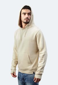 Felpa beige con tasca frontale, stringhe e polsini a coste. Realizzata in tessuto morbido, presenta una vestibilità rilassata e una texture liscia.