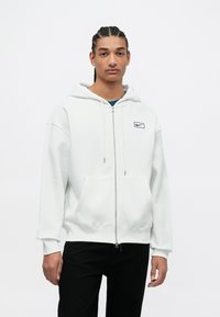 Biela mikina na zips vyrobená z mäkkého materiálu, s prednou klokankou a malým námorníckym logom Nike na ľavej strane hrude.
