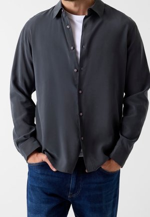 Man wearing a dark gray button-up shirt over a white t-shirt with hands in the pockets of blue jeans.