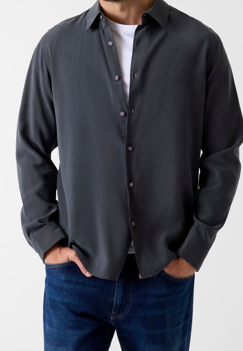 Homme portant une chemise gris foncé à boutons par-dessus un t-shirt blanc, les mains dans les poches d'un jean bleu.