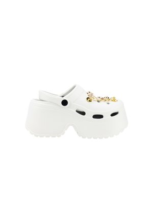 Zueco de plástico blanco con suela gruesa, agujeros de ventilación laterales y charms dorados decorativos en la parte superior. Superficie texturizada y forma redondeada.