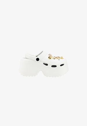 Zueco de plástico blanco con suela gruesa, agujeros de ventilación laterales y charms dorados decorativos en la parte superior. Superficie texturizada y forma redondeada.