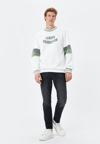 Sudadera blanca con el texto "LEADER GENERATION", rayas verdes y grises en las mangas, cuello redondo, combinada con jeans oscuros y zapatillas blancas.