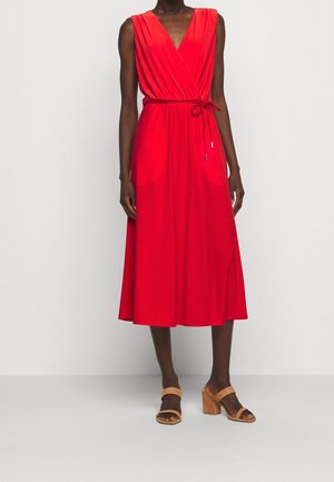 Vrouw draagt een mouwloze rode midi-jurk met V-hals en ceintuur in de taille, gecombineerd met sandalen met blokhak in beige, staand tegen een effen achtergrond.