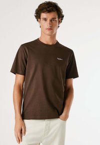 Brun bomulds t-shirt med korte ærmer, rund halsudskæring og et lille hvidt logo på venstre bryst. Enkel design, blød struktur.