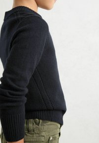 Maglione azzurro in maglia con collo e polsini a coste, caratterizzato da un dettaglio sulla cucitura laterale. Abbinato a pantaloni verde oliva.