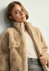Beżowy płaszcz z sztucznego futra z wysokim kołnierzem, zapięciem na zamek oraz kieszonką naszywaną. Miękka tekstura i lekko oversize'owy fason.