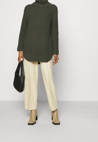 Tmavě zelený pletený svetr s vysokým rolákem, kombinovaný s krémovými kalhotami s širokými nohavicemi a béžovými kotníkov�ými botami na podpatku. Součástí je černá texturovaná kabelka.