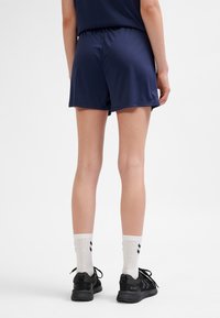 Persona que lleva pantalones cortos deportivos azul marino, calcetines blancos hasta el tobillo con cheurones negros y zapatos de correr negros, de pie contra un fondo blanco liso.