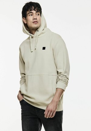 Sweatshirt à capuche beige clair avec un motif texturé, poche avant et un petit patch logo noir. Manches ajustées et une coupe décontractée.