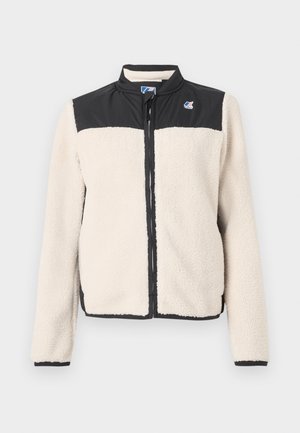 Beige fleecejack met zwarte panelen op de schouders, zwarte rits en logo-patch op de linkerborst, weergegeven op een witte achtergrond.