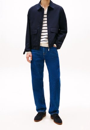 Veste bombardier navy avec deux poches avant, pull blanc avec des rayures navy, jeans en denim bleu et chaussures foncées.