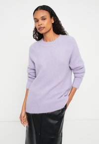 Kortsiriline naine, kes kannab lavendlitooni karvaseid sviitreid ja musta nahkseelikku, seisab ühe käega taskus valge tausta vastu.