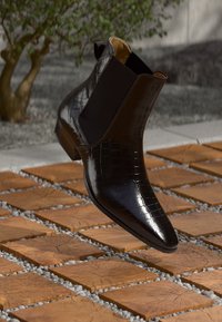 Bota de tornozelo em couro preto com textura em relevo de crocodilo, bico apontado, painéis laterais elásticos e um salto baixo, posicionada acima de uma superfície azulejada.