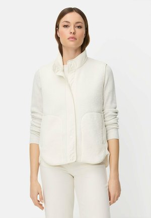 Kvinde iført en hvid ærmeløs fleecevest over en langærmet skjorte og hvide bukser, stående mod en ensfarvet hvid baggrund.