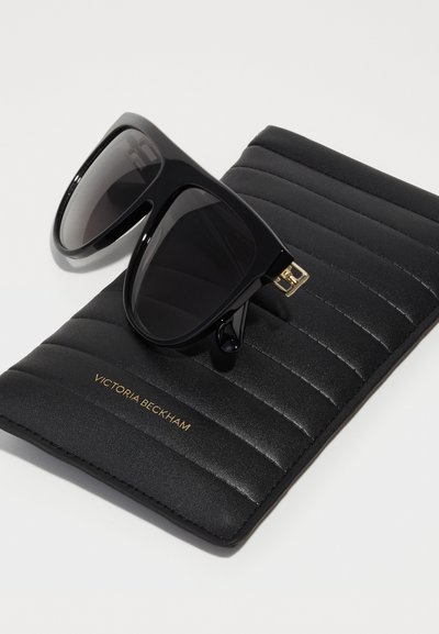 Óculos de sol oversized pretos com lentes escuras. Acompanhados por uma estojo preto texturizado com ranhuras horizontais e letras douradas.