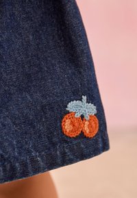 Denim fabric featuring an embroidered design of two orange fruits with a light blue leaf. Close-up of the hem detail.