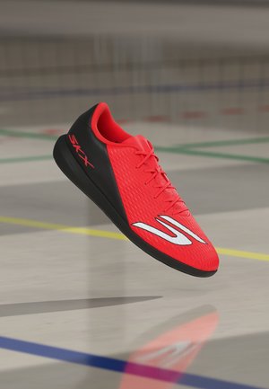 Roter und schwarzer Hallensportschuh mit Schnürsenkeln und weißem Logo, schwebt über einem Turnhallenboden mit farbigen Spielfeldlinien.