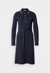 Tumši zils kokvilnas kleita ar garām piedurknēm, Polo apkakli, trim pogām un jostu viduklī. Izceļas ar baltu apdari.