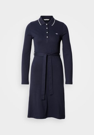 Granatowa sukienka z długim rękawem wykonana z bawełny, z kołnierzykiem polo, plisą na trzy guziki oraz dopasowanym paskiem w talii. Z ozdobnymi białymi wykończeniami.