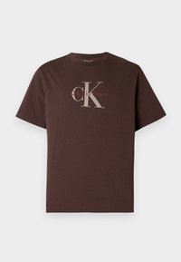 T-shirt de algodão castanha com mangas curtas, apresentando um grande logótipo cinzento 'CK' e texto vermelho mais pequeno 'Calvin Klein'. Design simples e de corte descontraído.