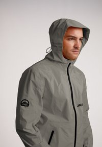 Giacca impermeabile grigia chiara con cappuccio, zip nera e logo sulla manica. Presenta una texture liscia e un cordino regolabile per il cappuccio.