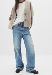 Femme portant un cardigan beige boutonné, un t-shirt blanc, un jean bleu à jambes larges et des mocassins noirs sur un fond uni.