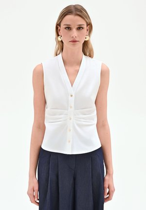 Femme portant un chemisier blanc sans manches boutonné avec un devant froncé et de grandes boucles d'oreilles dorées, associée à un pantalon plissé sombre, fond neutre.