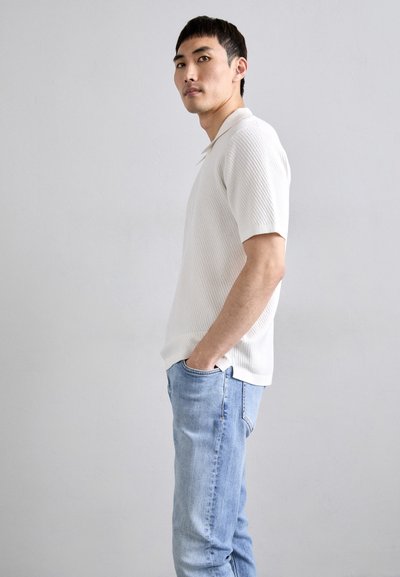 Witte geribbelde poloshirt met korte mouwen, relaxed fit met een kraag. In combinatie met lichtblauwe denim jeans. Effen achtergrond benadrukt de focus op het kledingstuk.