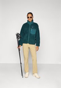 Giacca in pile color teal con tasca con zip, pantaloni beige e scarpe chiare. La persona tiene in mano dei bastoni da trekking mentre indossa occhiali da sole.