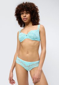 Γυναίκα με σγουρά μαλλιά που φοράει ελαφρύ μπλε δαντελένιο σουτιέν και ταιριαστό δαντελένιο εσώρουχο, στέκεται μπροστά σε ανοιχτό γκρι φόντο.