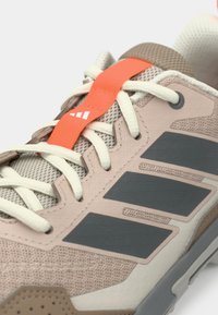 Μπεζ και γκρι αθλητικό παπούτσι με λευκά κορδόνια και πορτοκαλί λουρί με λευκό λογότυπο στην περιοχή της γλώσσας.