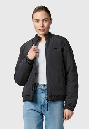 Vrouw draagt een zwarte bomberjack met rits over een wit shirt en lichtblauwe spijkerbroek, houdt de kraag van het jack vast met één hand.