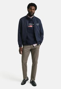 Navy bomberjack met een navy sweatshirt met USA en American flag details, gecombineerd met beige broek en zwarte schoenen.