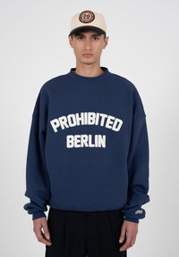 Jeune homme portant un sweat-shirt bleu marine avec le texte « PROHIBITED BERLIN » et une casquette beige avec un logo circulaire, debout devant un fond uni.