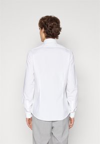 Persona con cabello rizado corto que lleva una camiseta blanca de manga larga ajustada y pantalones claros grises, de espaldas contra un fondo liso.