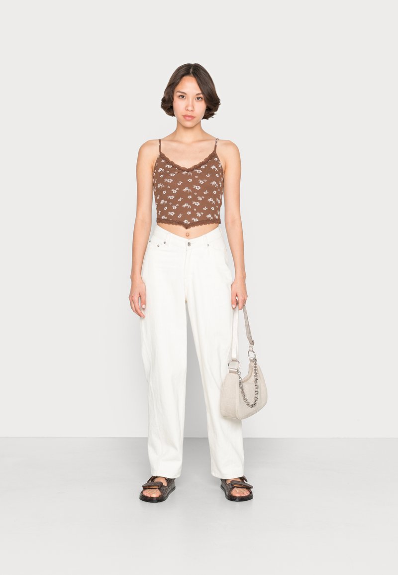 Top corto marrone con pizzo e motivo floreale, abbinato a pantaloni bianchi larghi e sandali neri texturizzati. Tiene in mano una piccola borsa beige.