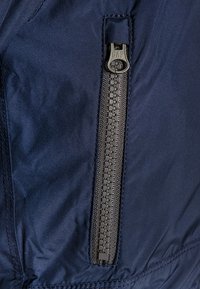 Granatowy, lekki materiał o gładkiej fakturze z wyraźnym czarnym zamkiem błyskawicznym. Zamek ma kwadratowy uchwyt i ozdobne przeszycia.