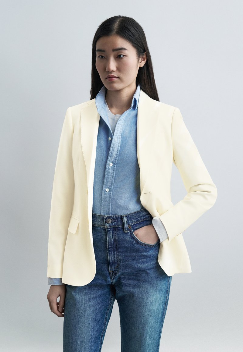 Νεαρή γυναίκα με μακριά σκούρα μαλλιά, ντυμένη μ ε μπεζ σακάκι πάνω από ανοιχτό μπλε πουκάμισο και μπλε τζιν, με το ένα χέρι στην τσέπη, ουδέτερο φόντο.