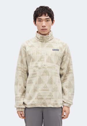 Junger Mann trägt einen beige gemusterten Fleece-Pullover mit Viertelreißverschluss und Columbia-Logo auf der Brust, steht vor einem einfarbigen Hintergrund.