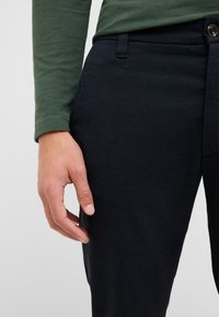 Dunkelblaue Hose aus strukturiertem Stoff mit Seitentasche, Knopfverschluss und grünem Langarmhemd. Hand ruht auf dem Oberschenkel.