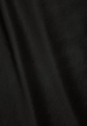 Femme portant une longue robe noire avec des manches bouffantes et des détails en dentelle, debout de profil sur un fond gris clair uni.