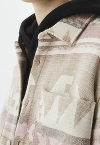 Camicia tessuta in tonalità beige e rosa con motivi geometrici, dotata di una tasca sul petto e colletto a punta, indossata sopra un felpa nera con cappuccio.