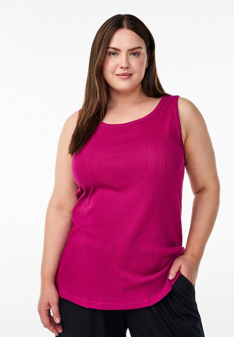 Una donna con lunghi capelli castani indossa un top aderente senza maniche di un vivace colore rosa e pantaloni neri, posando su uno sfondo bianco semplice.