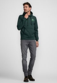 Sudadera verde oscuro con un bolsillo frontal y capucha con cordón, combinada con jeans gris ajustados y botas negras con cordones. Diseño minimalista.