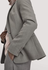 Grijze op maat gemaakte blazer van textuurstof, met drie knopen op de manchetten en een gestructureerde revers, gecombineerd met een roze trui eronder.