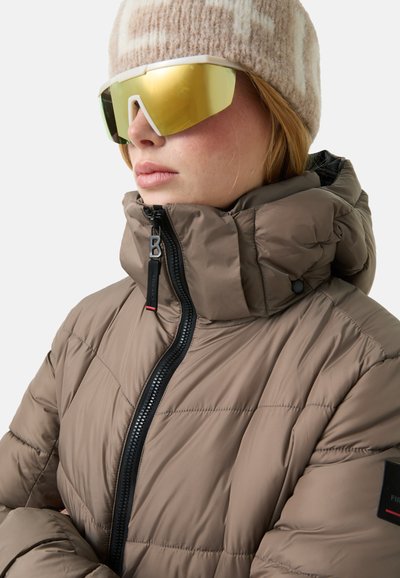 Gepolsterte braune Jacke mit hohem Kragen, Reißverschluss und strukturierter Oberfläche. Zubehör umfasst eine beigefarbene gestrickte Mütze und große reflektierende Sonnenbrillen.