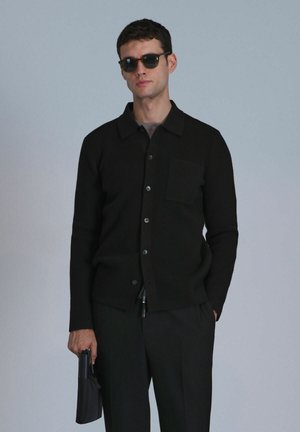 Schwarze gerippte Strickjacke mit Kragen, Vorderknöpfen und einer Brusttasche. Das Model hält ein schwarzes Accessoire und trägt eine dunkle Sonnenbrille.